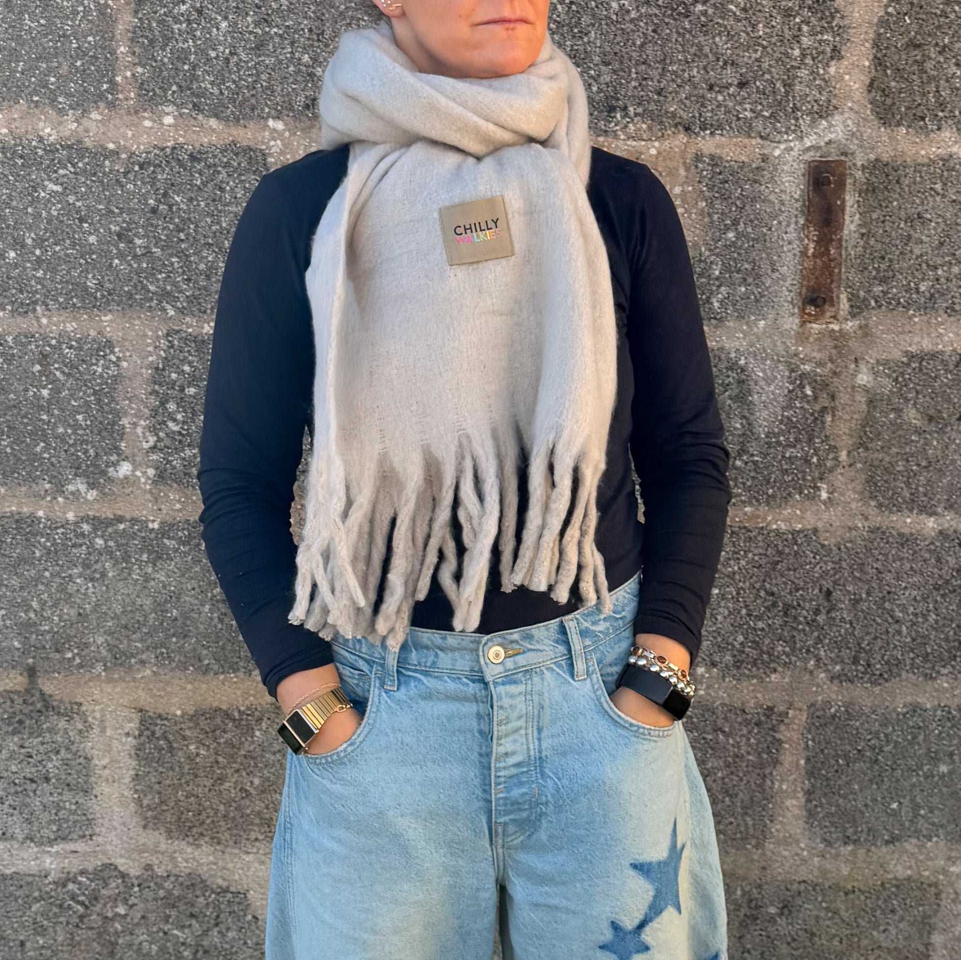 
A person wearing an oversized light beige-coloured Chilly WALKIES scarf with tasselled ends, styled over a black top and denim jeans.